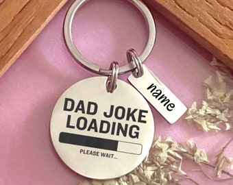Op de afbeelding: Zilveren sleutelhanger met een rond label waarop "Dad Joke Loading" staat met een voortgangsbalk en de tekst "Please Wait..." eronder. Een kleiner label met het woord "name" is aan de sleutelhanger bevestigd.