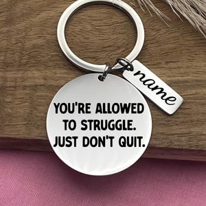 Puede incluir: Un llavero plateado con un colgante redondo y una etiqueta rectangular. El colgante redondo tiene el texto "YOU'RE ALLOWED TO STRUGGLE. JUST DON'T QUIT." La etiqueta rectangular tiene la palabra "name" en cursiva.