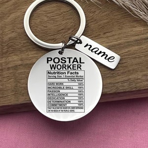 Könnte beinhalten: Ein silberner Schlüsselanhänger mit einem runden Anhänger, auf dem "POSTAL WORKER" steht und "Nutrition Facts" mit einem prozentualen Tageswert für harte Arbeit, unglaubliche Fähigkeiten, Leidenschaft, Intelligenz, Hingabe, Entschlossenheit und Engagement aufgelistet sind. Der Anhänger enthält auch den Text "Der Tageswert kann je nach Bedarf der Menschen, die ich bediene, höher oder niedriger sein". Ein kleinerer silberner Anhänger mit dem Wort "name" hängt am Schlüsselanhänger.