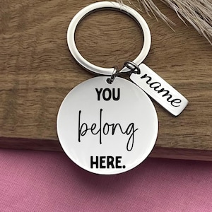 May include: A silver keychain with a round pendant that reads "YOU belong HERE." A smaller rectangular tag is attached, with the word "name" engraved. The keychain is on a wooden surface.