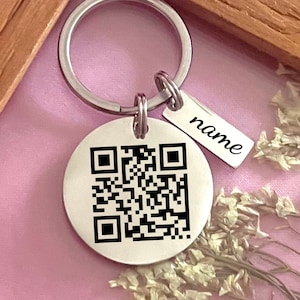 Peut inclure: Un porte-clés argenté avec une étiquette ronde comportant un code QR noir et blanc. L'étiquette porte également le mot "name" gravé.
