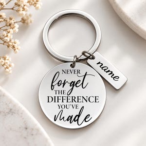 Può includere: Un portachiavi in metallo color argento con un ciondolo circolare inciso con il testo nero NEVER forget THE DIFFERENCE YOU'VE made e una targhetta rettangolare più piccola incisa con la parola name, fissato a un anello.