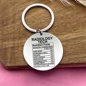 May include: Silver keychain with a round tag that reads "RADIOLOGY TECH" and lists "Nutrition Facts" with a serving size of "1 Essential Worker". The tag lists qualities like "Hard Work", "Incredible Skill", "Passion", "Intelligence", "Dedication", "Determination", and "Commitment", all with a 100% daily value.