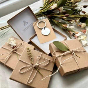 May include: Four small brown boxes with twine bows and small floral accents. Each box contains a small gift. A silver key chain with a round tag is also pictured.