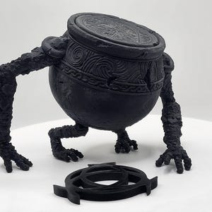 May include: A black, stylized sculpture of a pot-like creature with arms and legs, and a separate, matching emblem. The pot has detailed carvings and a lid. The sculpture is on a white surface.