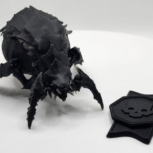 May include: A black, 3D-printed insect-like creature with multiple legs and a spiky carapace. Two black, geometric tokens with a skull emblem are also present. The objects are on a white surface.