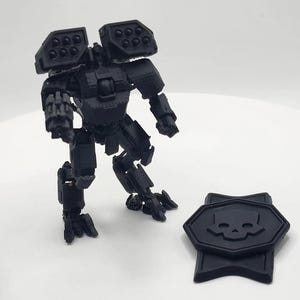 May include: A black, articulated robot model with missile launchers on its shoulders and a detailed design. A black emblem with a skull is next to the robot. The model is on a white surface.