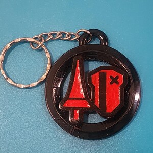 May include: Black keychain with a circular pendant featuring a red and black design. The design includes a red spear and a red shield with a black X. The keychain has a silver key ring and chain.