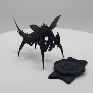 May include: A black, 3D printed, insect-like creature with wings and multiple legs. The creature is standing on a white surface. A black, skull-shaped token is in the foreground.