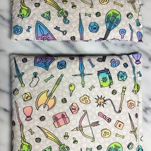 May include: Two grey fabric eye pillows with a rainbow-coloured pattern of dice, swords, keys, and other fantasy-themed items. The larger pillow is 12 inches long and the smaller pillow is 6 inches long.