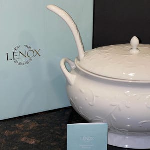 May include: White Lenox brand soup tureen with a lid and a ladle. The tureen has a floral design and is sitting on a brown surface. The tureen is in a box with the Lenox logo on it.