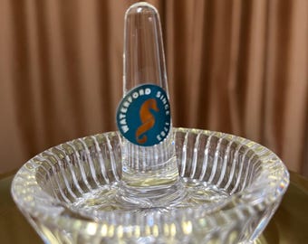 May include: A clear glass ring holder with a raised center post and a decorative, fluted base. The holder has a small, round sticker on the post that says "Waterford Since 1825" and features a blue seahorse.