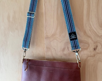 May include: A brown leather crossbody bag with a blue and white patterned strap. The bag has a zippered top and a small pocket on the front. The strap is adjustable and attaches to the bag with silver-colored hardware. The bag is hanging against a light brown wooden background.