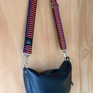 May include: Black leather crossbody bag with a patterned strap in orange, purple, and black. The strap is attached to the bag with silver-colored hardware. The bag has a curved shape and a flap closure. The brand name "LO HAZEL" is visible on the strap.