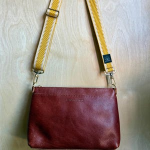 May include: A dark red leather crossbody bag with a textured surface and a zipper closure. The bag features a detachable, adjustable strap in mustard yellow with white chevron patterns and a small black tag labeled LO & HAZEL.