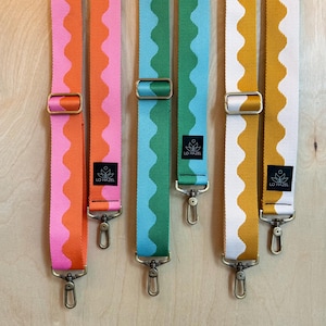 May include: Three adjustable bag straps featuring a wavy pattern. The first strap is orange and pink, the second is light blue and green, and the third is white and mustard yellow. Each strap includes metal hardware and a black label reading LO HAZEL.