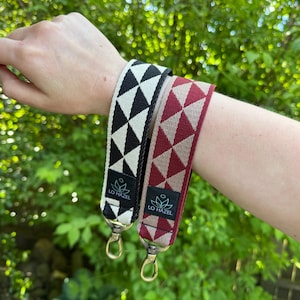 May include: Two patterned wrist straps with gold-colored clasps. One strap features a black and white triangle design, while the other has a burgundy and beige triangle pattern. Both straps have a black label with the text "LO HAZEL".