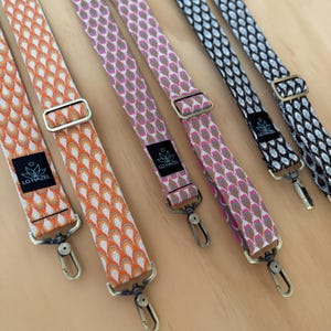 May include: A collection of patterned bag straps in various colors, including orange, pink, and black. Each strap features a repeating geometric design and is equipped with metal hardware, including adjustable buckles and swivel clasps for easy attachment.