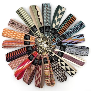 May include: A collection of colorful woven wrist keychains with various geometric and patterned designs. Each keychain features a metal clasp and a black tag with the text "LO HAZE". The keychains are arranged in a circular pattern.