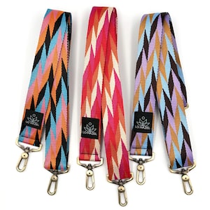 May include: Three colorful patterned bag straps with gold-colored hardware. The straps feature geometric designs in various color combinations, including orange, blue, pink, and brown. Each strap has a black label with the text "LO HAZEL".