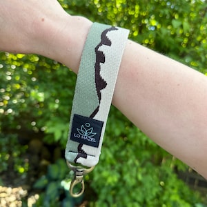 May include: A green and brown patterned wristlet with a gold-colored clasp. The wristlet has a black tag with the text "LO HAZEL" and a floral design. The wristlet is made of woven fabric.