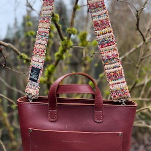 May include: A burgundy leather tote bag with a colorful, textured strap. The strap is woven with threads of red, yellow, blue, and white. The bag has a small front pocket and two short handles. The brand logo "LO HAZEL" is visible on the strap.
