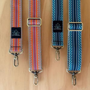 May include: Four patterned bag straps in orange, purple, and blue with gold-colored hardware. Each strap has a black label with the text "LO HAZEL" and a floral design. The straps are displayed on a light brown wooden surface.