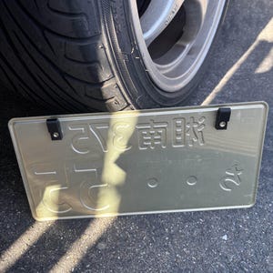 May include: A rectangular, light-colored license plate with Japanese characters and the number 24. The plate is held by two black straps. A car tire and wheel are visible in the background.