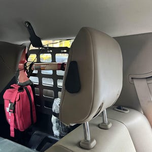 May include: Interior car view with a tan headrest and seat. A black metal cargo barrier is behind the seat, holding a red first aid kit and other items. The car's interior is neutral-toned.