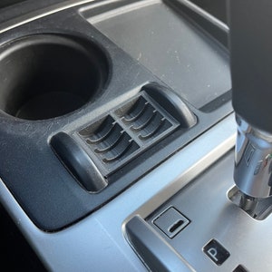 May include: A close-up of a car's interior, showing a black cup holder, a silver gear shift lever, and a black plastic vent with a silver trim. The gear shift lever has the letters "P", "R", and "N" printed on it.