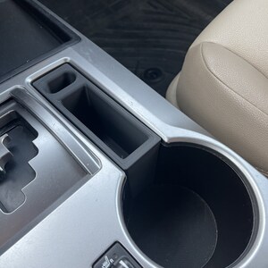 May include: A close-up of the interior of a car, showing a silver dashboard with a cup holder and a small storage compartment. The cup holder is empty and the storage compartment is closed.