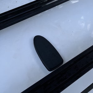 以下が含まれることがあります： 黒い楕円形の物体、おそらく車の部品が白い表面に置かれています。 物体は滑らかで、固い素材でできているようです。 黒い長方形のレールが白い表面を縁取っています。