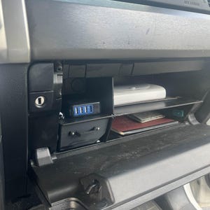 May include: A black car's glove compartment is open, revealing a black storage compartment with a USB charging port and a red and black notebook. The compartment is lined with a gray fabric.
