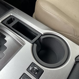 Op de afbeelding: Een close-up van de middenconsole van een auto met een bekerhouder, twee zwarte knoppen met zilveren knoppen en een klein opbergvak. De console is gemaakt van zilverkleurig metaal.