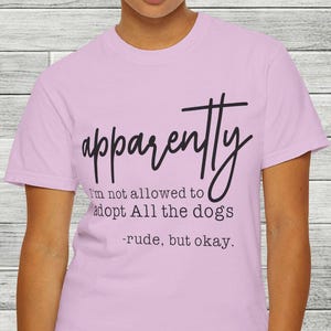 Può includere: Una maglietta lilla con la parola "apparently" in un carattere corsivo. Sotto la parola, il testo recita: "I'm not allowed to adopt All the dogs - rude, but okay."