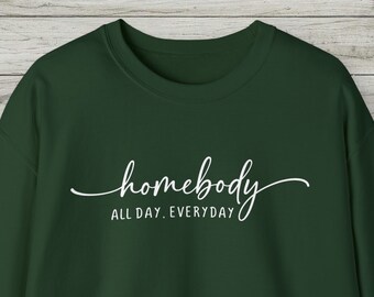 May include: A dark forest green crewneck sweatshirt featuring the white text "homebody" in a cursive script font, with the words "ALL DAY. EVERYDAY" printed in a simple sans-serif font directly underneath the main script on the chest area.
