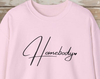 May include: A light pink crewneck sweatshirt featuring the word Homebody written in a black cursive script font across the chest, with a small black heart symbol at the end of the text. The garment has long sleeves and a ribbed collar.