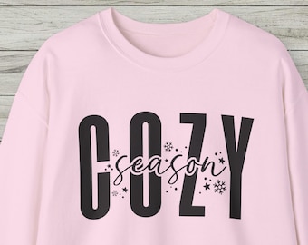 May include: A light pink long-sleeve sweatshirt featuring the black text COZY in a bold, sans-serif font. The word season is written in a smaller, black cursive script across the center, surrounded by small black snowflake and star icons.