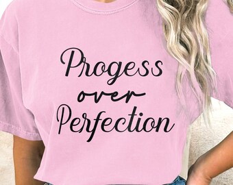 May include: A pale pink t-shirt featuring the phrase "Progress over Perfection" in black cursive lettering. The top has a round neck and short sleeves. The material looks soft and comfortable.