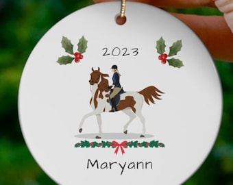 Könnte beinhalten: Ein rundes, weißes Keramikornament mit einem braun-weißen Scheckenpferd und einer reitenden Person in Reitkleidung. Das Design zeigt den Text 2023 und Maryann, verziert mit grünen Stechpalmenblättern, roten Beeren und einer grünen Girlande mit roter Schleife.