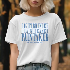 May include: White t-shirt with blue text that reads "LIGHTBRINGER, CHAINBREAKER, PAINTAKER" and "HE WILL NEVER FAIL". The shirt is being worn by a person with short blonde hair. The background is blurred.