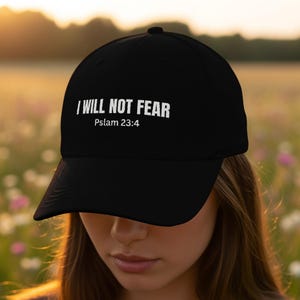 May include: Black baseball cap featuring the white text "I WILL NOT FEAR" and "Psalm 23:4". The cap has a curved peak and a button on the crown. The background is a blurred field of flowers.