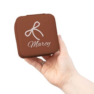 Peut inclure: Une boîte à bijoux carrée marron avec un motif de nœud en perles blanches et le nom "Marcy" en écriture cursive. La boîte est tenue dans une main, mettant en valeur sa taille compacte et son détail personnalisé. Le design est simple et élégant.