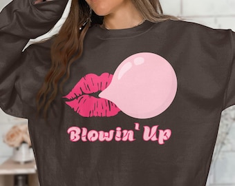 Può includere: Una felpa marrone con una grafica di chewing gum rosa e labbra rosa. Il testo "Blowin' Up" è stampato in rosa sotto la grafica.