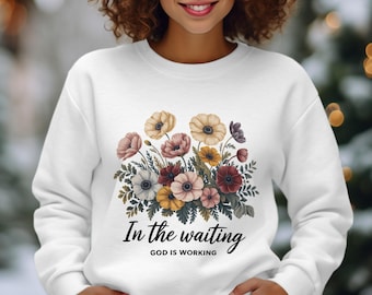 May include: White sweatshirt featuring a floral design with various colors of anemones and greenery. The text "In the waiting GOD IS WORKING" is printed below the flowers. The sweatshirt is a crew neck style.