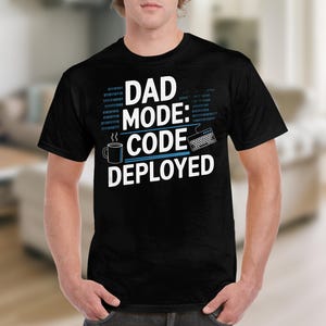 Może przedstawiać: Czarny T-shirt z białym tekstem, który brzmi "Dad Mode: Code Deployed". Tekst jest otoczony niebieskim wzorem przypominającym kod. Wzór zawiera również mały obrazek filiżanki kawy i klawiatury komputerowej.