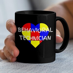 May include: Black ceramic mug featuring a heart-shaped design in red, yellow, and blue. The words "BEHAVIORAL TECHNICIAN" are printed in white across the heart. The mug has a curved handle.