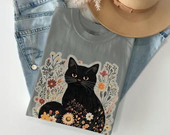 Può includere: Una maglietta grigia con un'illustrazione di un gatto nero circondato da fiori colorati. Il gatto ha gli occhi gialli e una lunga coda nera. La maglietta è piegata sopra jeans blu e un cappello beige.