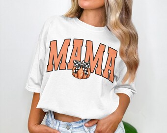 Pode incluir: Camiseta branca com a palavra "MAMA" em letras laranja. Uma abóbora com um laço xadrez preto e branco está centrada abaixo da palavra. A camisa é de estilo casual.