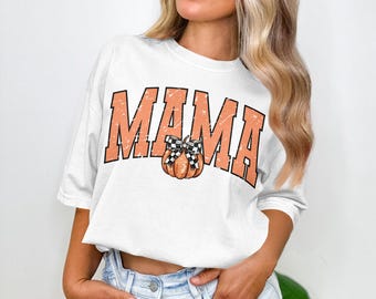 Pode incluir: Camiseta branca com a palavra "MAMA" em letras de bloco laranja. Uma abóbora com um laço xadrez preto e branco está centrada sob a palavra "MAMA". A camisa é de estilo casual.